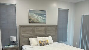 Een slaapkamer met een houten bed, een nachtkastje, een lamp, een schilderij en een raam met jaloezieën.