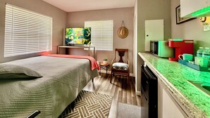 A compact hotel room with a bed, a TV, a chair, a small table, and a kitchenette.