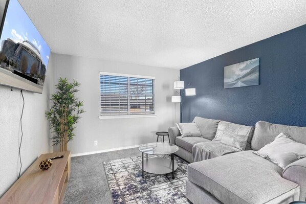 A modern living room with a grey sofa, a coffee table, a potted plant, and a TV mounted on the wall. - Arlington 1BR | AT&T, Globe Life, 6 Flags, Parking (Arlington)
