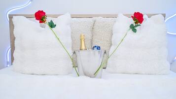 A bed with fluffy white pillows, a champagne bottle, and two red roses.