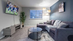 Ein modernes Wohnzimmer mit grauem Sofa, einem Glastisch, einem an der Wand montierten Fernseher, einer Topfpflanze und einem Fenster mit Jalousien.