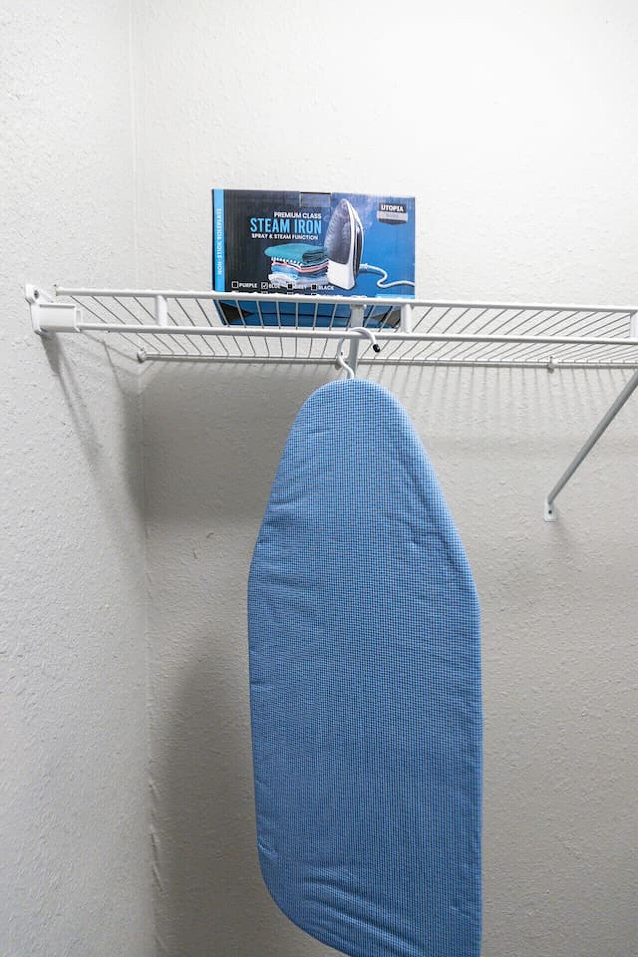 Ein blaues Bügelbrett hängt an einem Metallgestell vor einer weißen Wand.