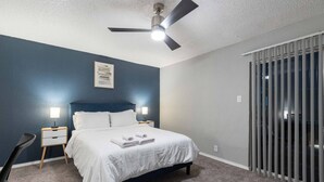 A bedroom with a bed, two bedside tables with lamps, a ceiling fan, a window with blinds, and a door with a vertical handle. - Arlington 1BR | AT&T, Globe Life, 6 Flags, Parking (Arlington)