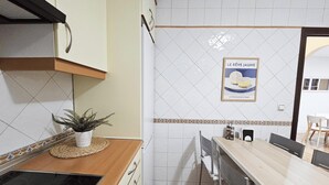Eine Küche mit weißen Fliesen, einem gerahmten Plakat an der Wand, einem Blumentopf auf der Arbeitsfläche und einem Essbereich, der durch einen offenen Durchgang sichtbar ist.