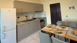 A modern kitchen with grey cabinets, a white refrigerator, and a dining area set for four with a wooden table and grey chairs. - Cozy Modern Apartment with Sea View (Sarandë)