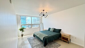 A bedroom with a large window, a bed with green bedding, a floor lamp, and a small wooden nightstand. - Spacious Midtown Miami 2-Bedroom Suite with Bay View (Miami)