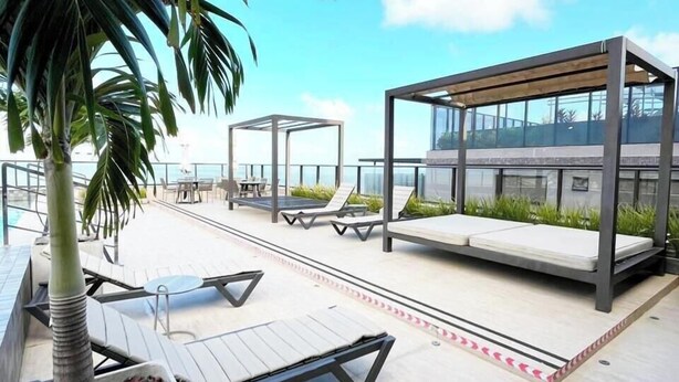 A rooftop terrace with sun loungers, a palm tree, and a glass-enclosed seating area. - Csq Jardins By DayByDay (Joao Pessoa)