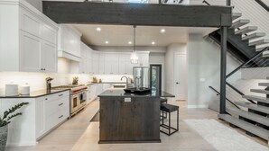 A modern kitchen with white cabinets, a central island, and a staircase with a metal railing. - Modern Mountain Getaway Near Park City | Sleeps 15 (Park City)