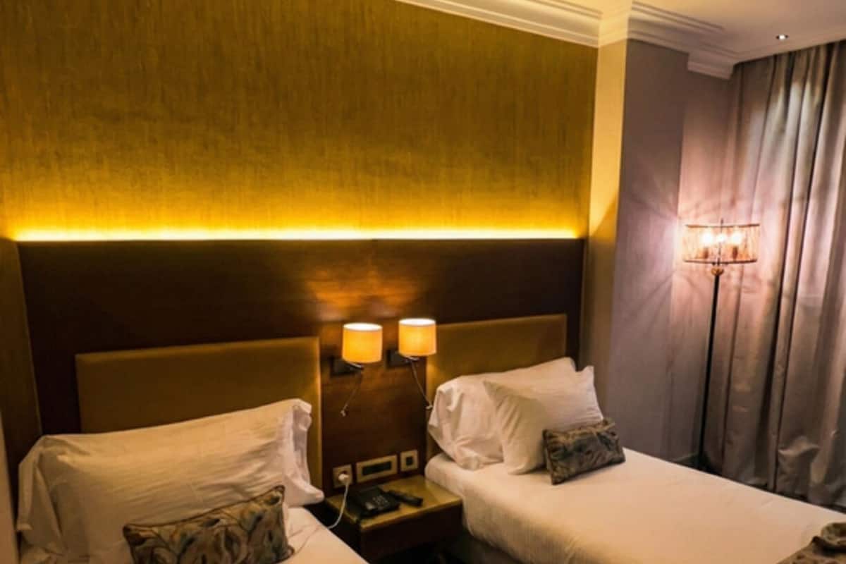 A hotel room with two beds, a headboard with integrated lighting, a bedside table with a lamp, and a patterned carpet.