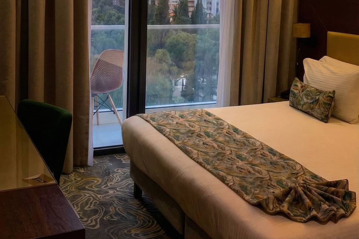A hotel room with a large bed, a desk, and a view of the cityscape through the window.