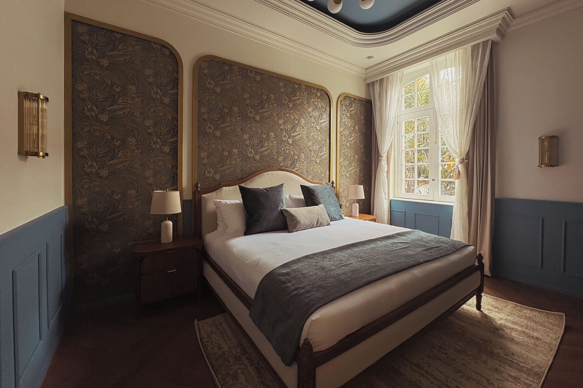 A bedroom with a large bed, two bedside tables with lamps, a window with curtains, and a decorative ceiling light.