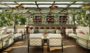 Un espace extérieur aménagé avec des meubles de jardin, comprenant des canapés et des tables à rayures, un bar avec des étagères remplies de bouteilles, et une vue sur les arbres.