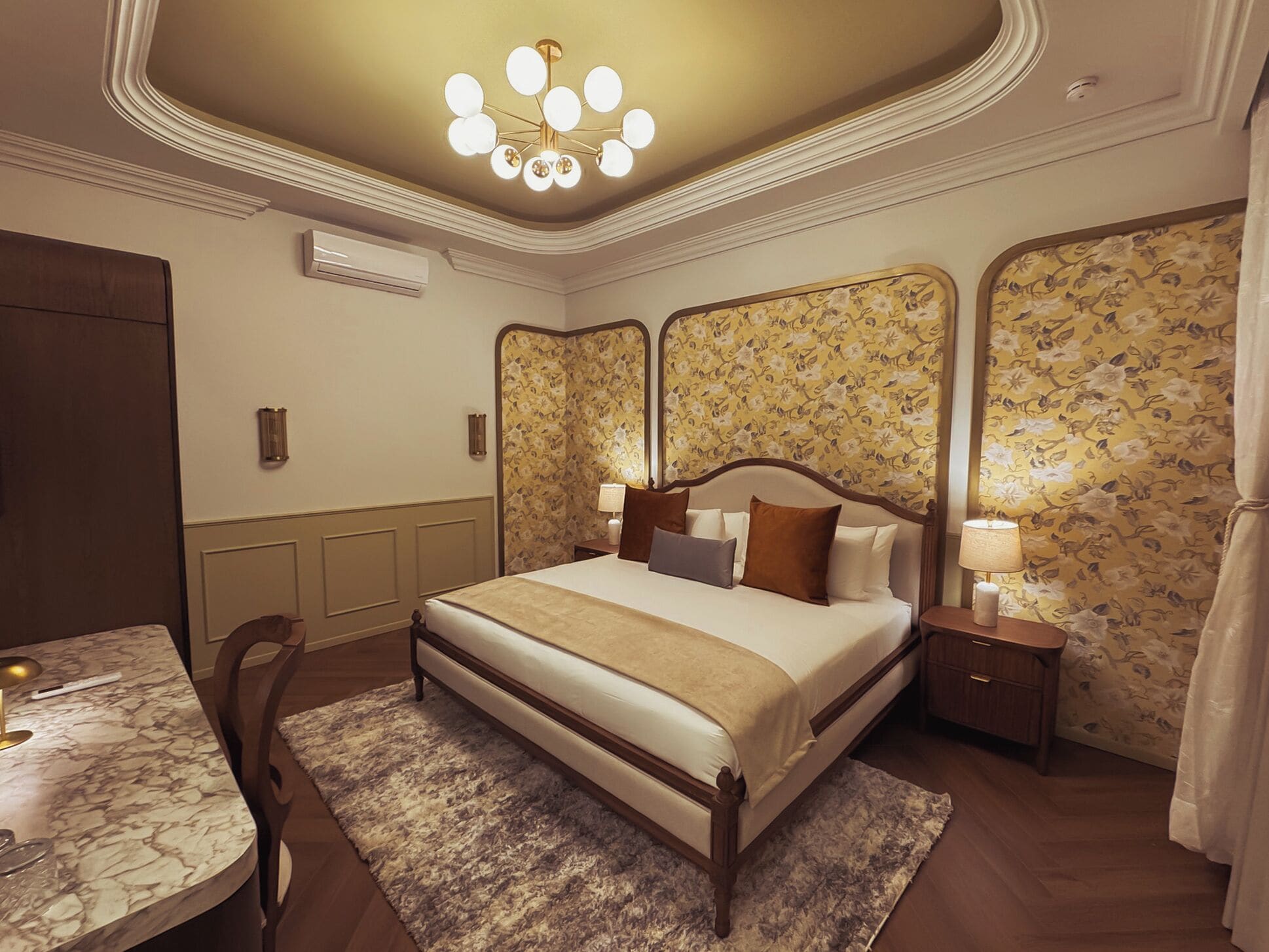 Une chambre d’hôtel avec un grand lit, un bureau avec une lampe, une table de chevet avec une lampe et un papier peint à motifs floraux.