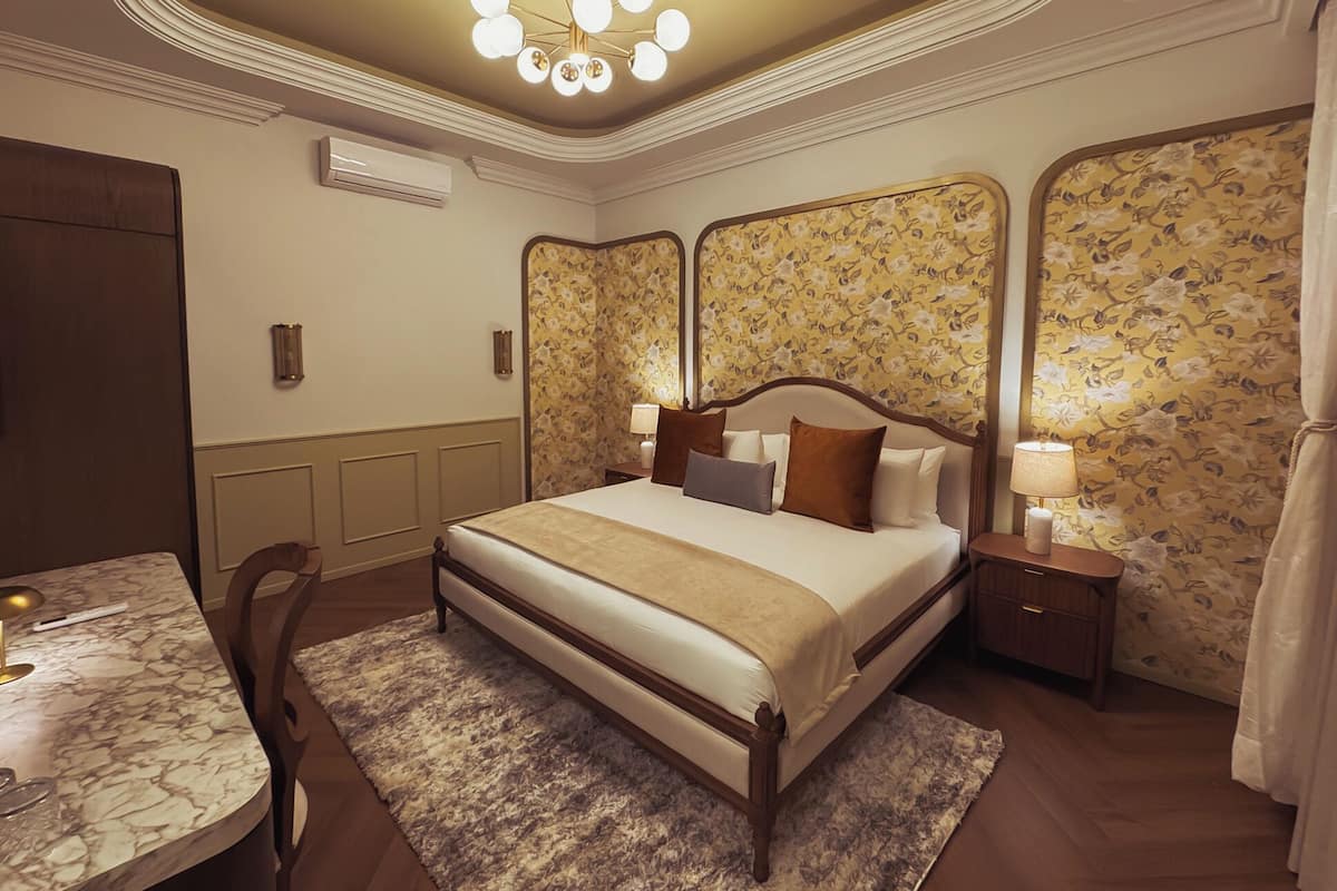 A hotel room with a large bed, a desk with a lamp, a nightstand with a lamp, and floral wallpaper.