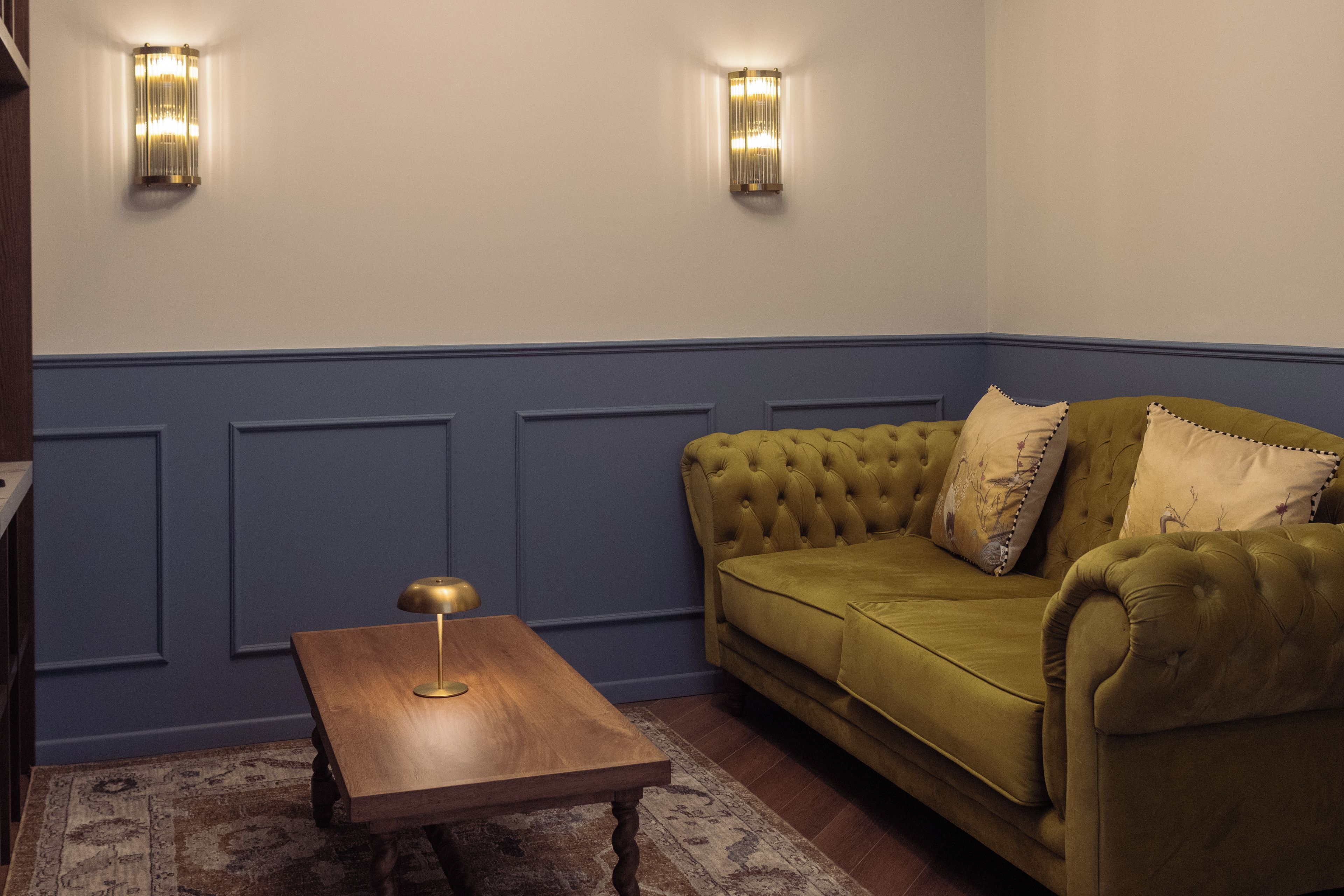 Una habitación con un sofá verde, una mesa de madera y dos lámparas fijadas a la pared.