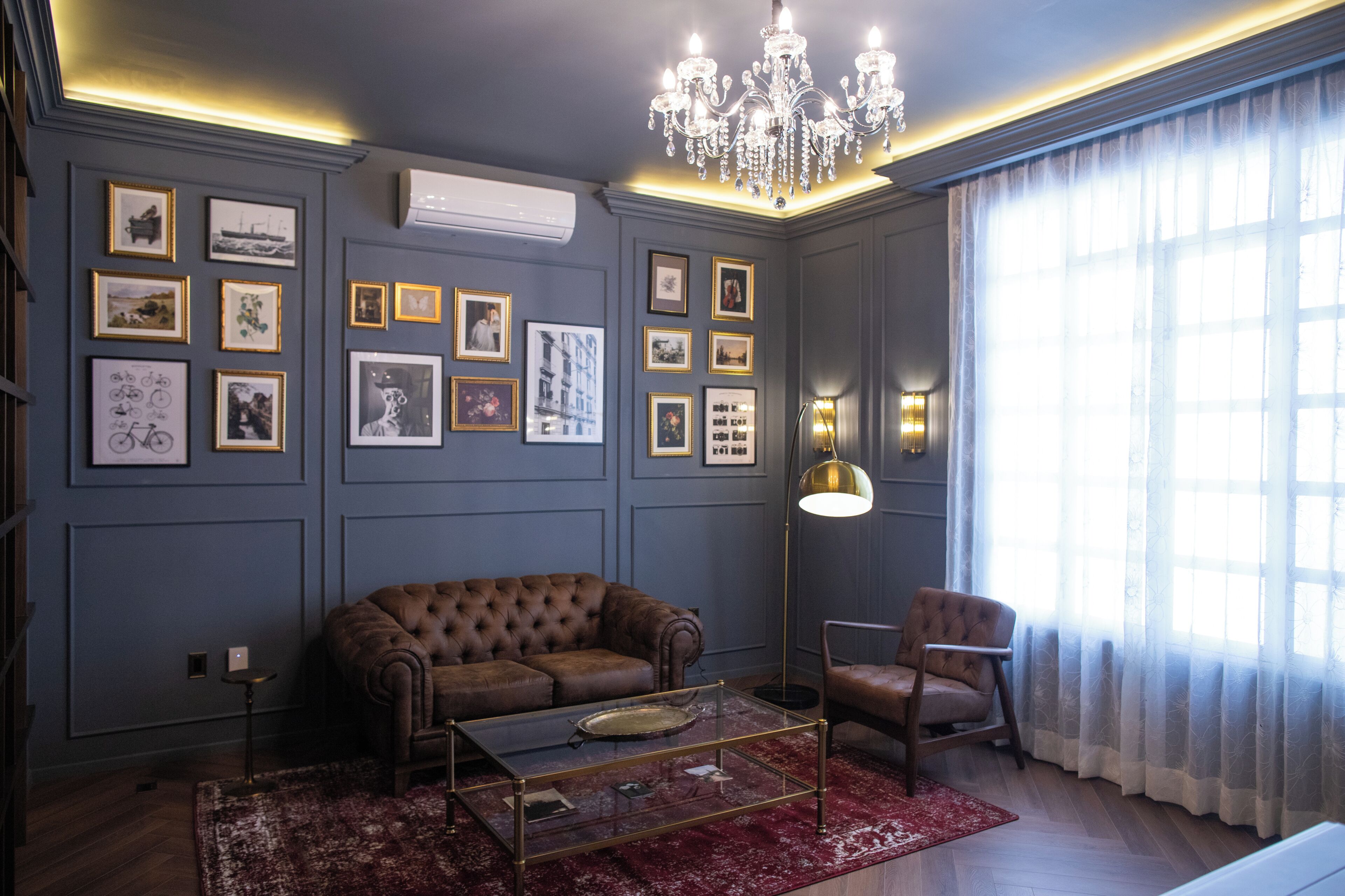 A room with a brown sofa, a glass coffee table, a red rug, a chandelier, framed pictures on the wall, and a large window with sheer curtains.