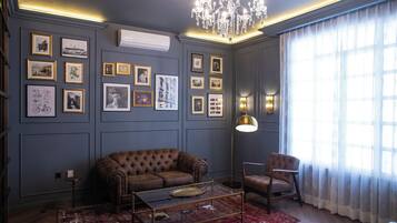 A room with a brown sofa, a glass coffee table, a red rug, a chandelier, framed pictures on the wall, and a large window with sheer curtains.