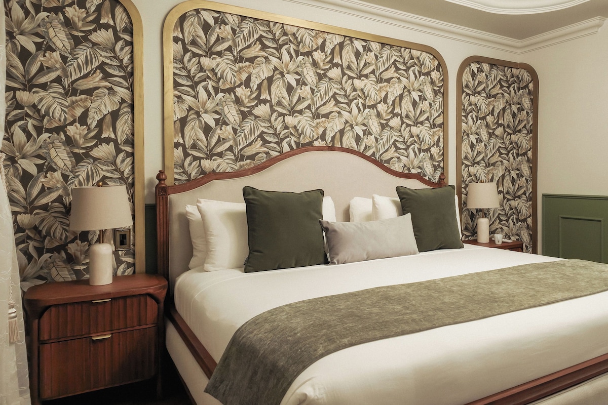 A neatly made bed with a wooden headboard, flanked by two bedside tables with lamps, in a room with floral wallpaper and a green accent wall.