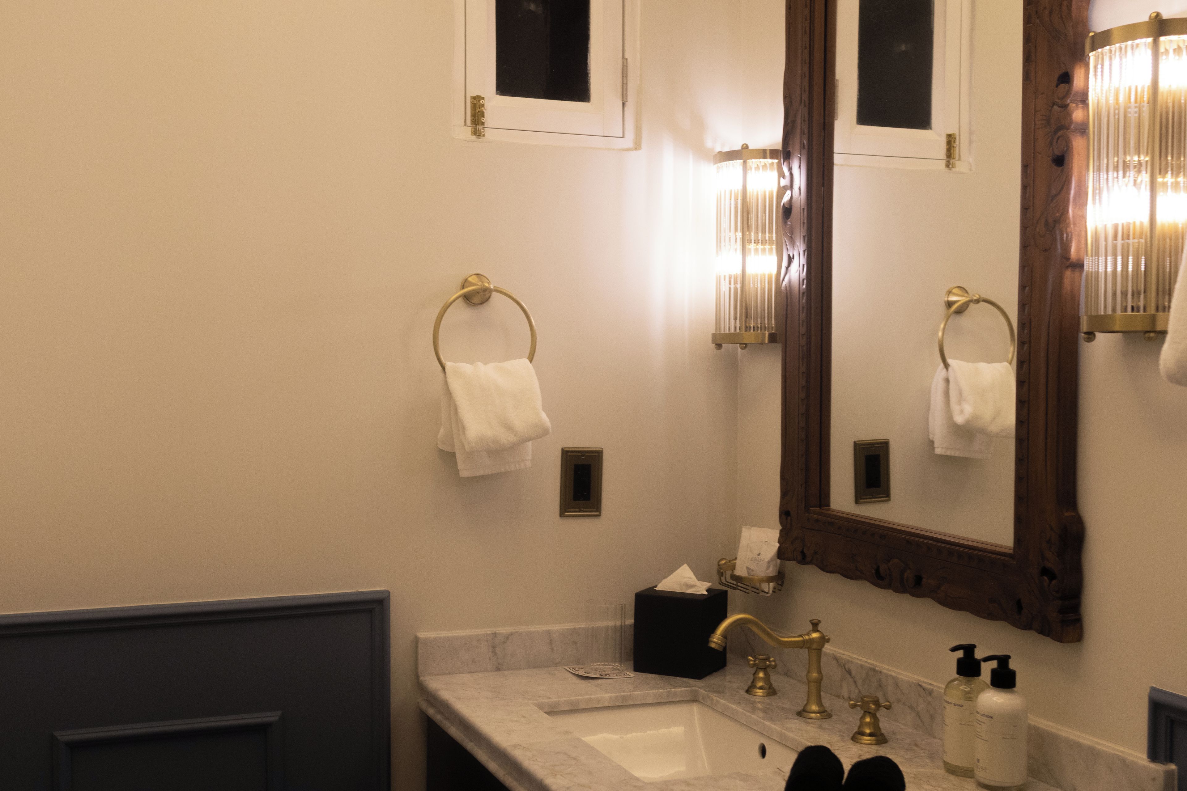 A bathroom with a marble sink, gold faucets, a large mirror, and two towel rings.