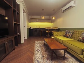Un espacio habitable compacto con un sofá, una mesa de centro, un televisor, una zona de cocina y una alfombra estampada.