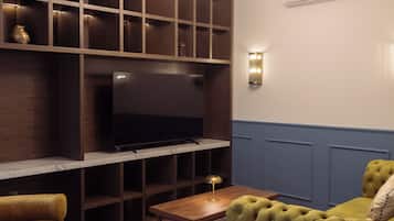 Una sala de estar con una televisión sobre un mueble de madera, una mesita, un sofá verde y una lámpara fijada a la pared.
