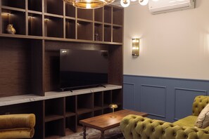 A living room with a television on a wooden stand, a small table, a green sofa, and a wall-mounted light fixture. - Roma Nueve Dos Hotel Boutique (Mexico City)
