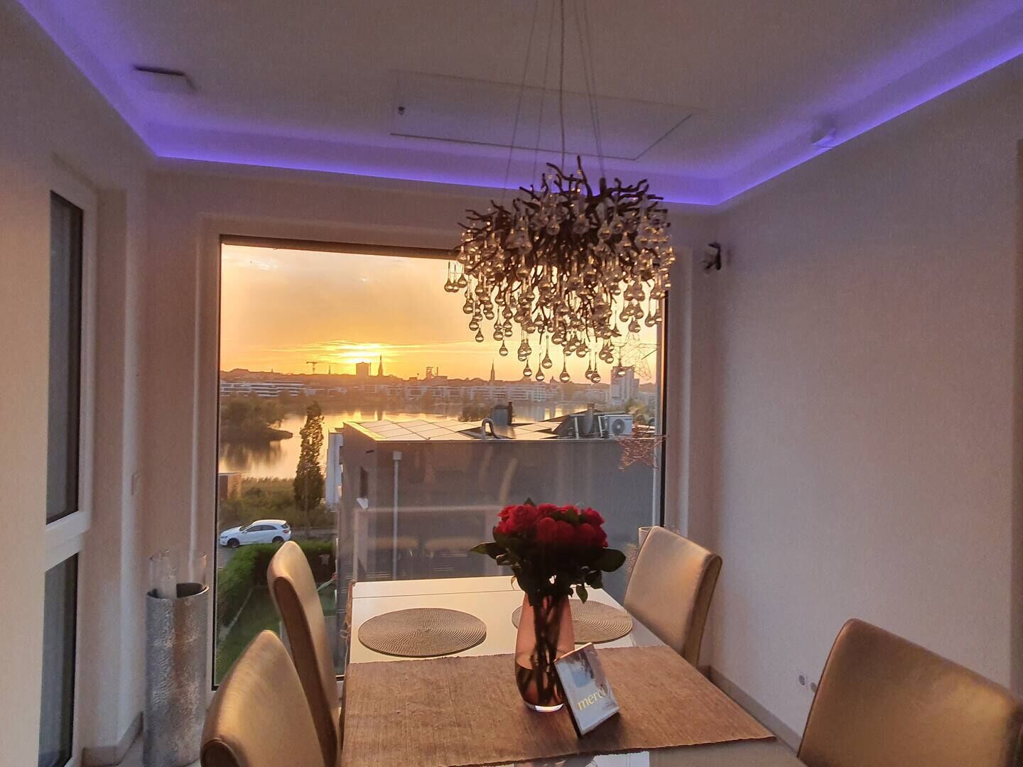 A dining area with a table set for two, a vase with red flowers, and a view of a sunset over a cityscape.