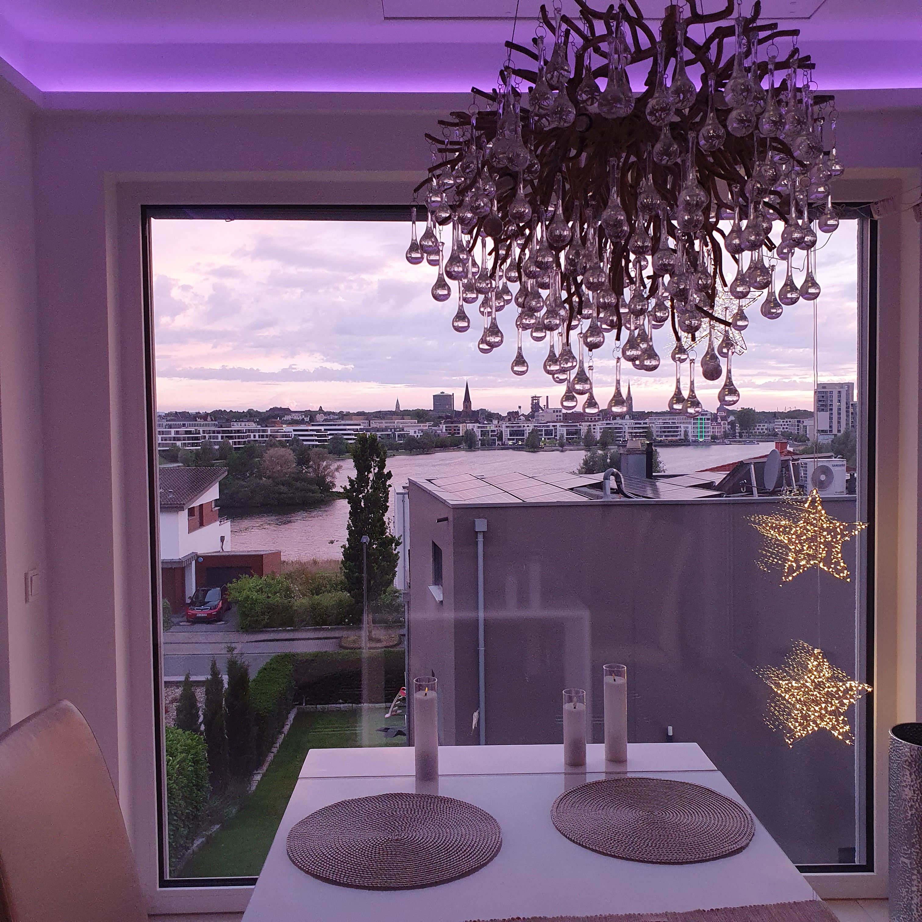 Ein Zimmer mit großem Fenster und Blick auf eine Stadtlandschaft, einer Kristallleuchtre und einem Tisch mit Unterlagen.