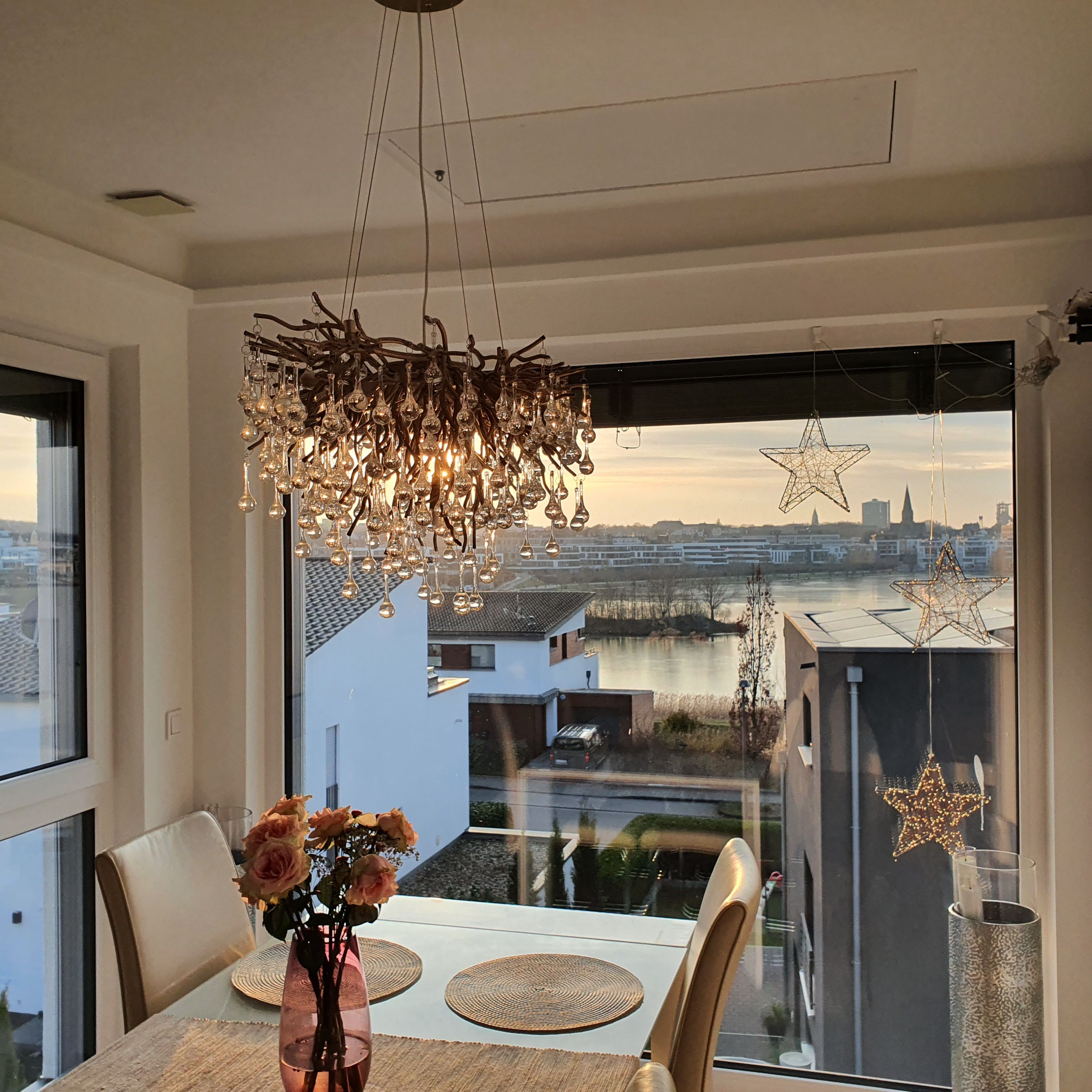 Ein Essbereich mit Tisch, Stühlen, einer Vase mit Blumen, einer Hängelampe und Blick auf eine Stadtkulisse durch ein Fenster.