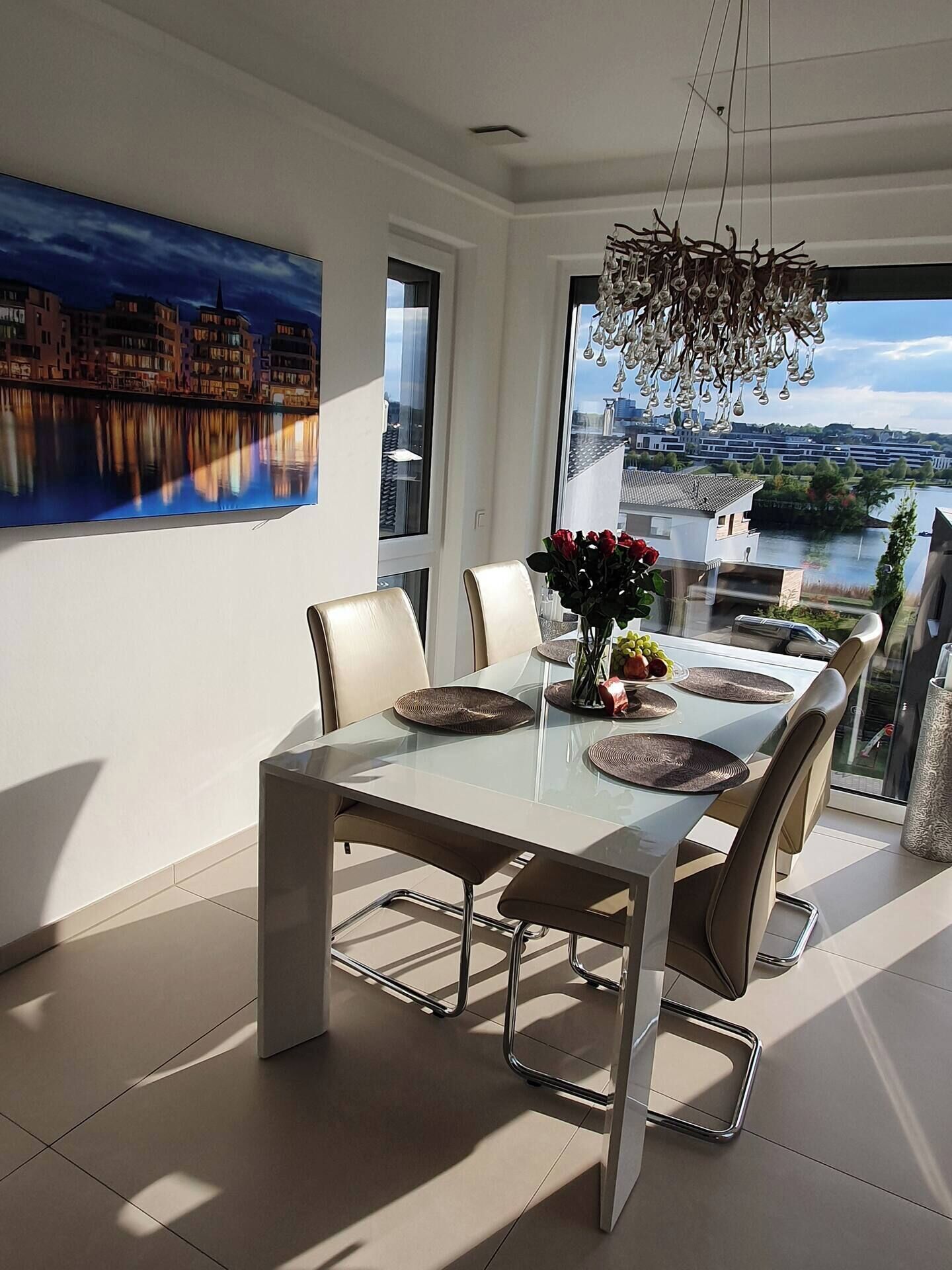 A modern dining area with a glass table, metal chairs, and a view of a river and buildings.