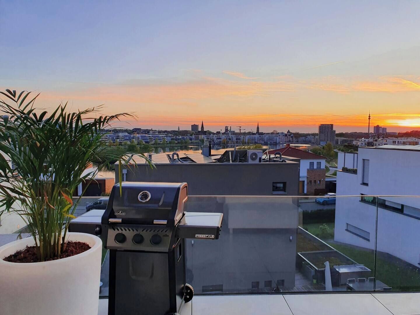 Une terrasse sur le toit, avec un barbecue, un pot de fleurs et une vue sur les toits de la ville au coucher du soleil.
