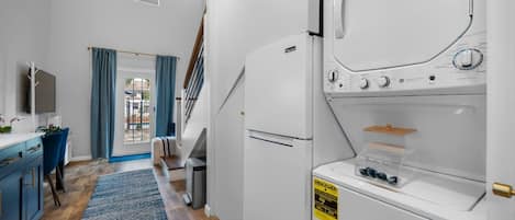 Une petite buanderie comprenant un lave-linge et un sèche-linge, un petit bureau avec une chaise, et une vue sur un espace extérieur accessible par une porte.