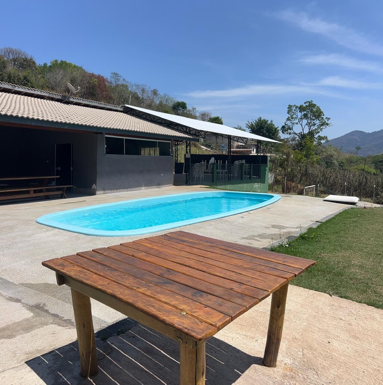 A swimming pool with a wooden table in the foreground, a covered patio area, and a view of a house with a tiled roof and a fenced yard.