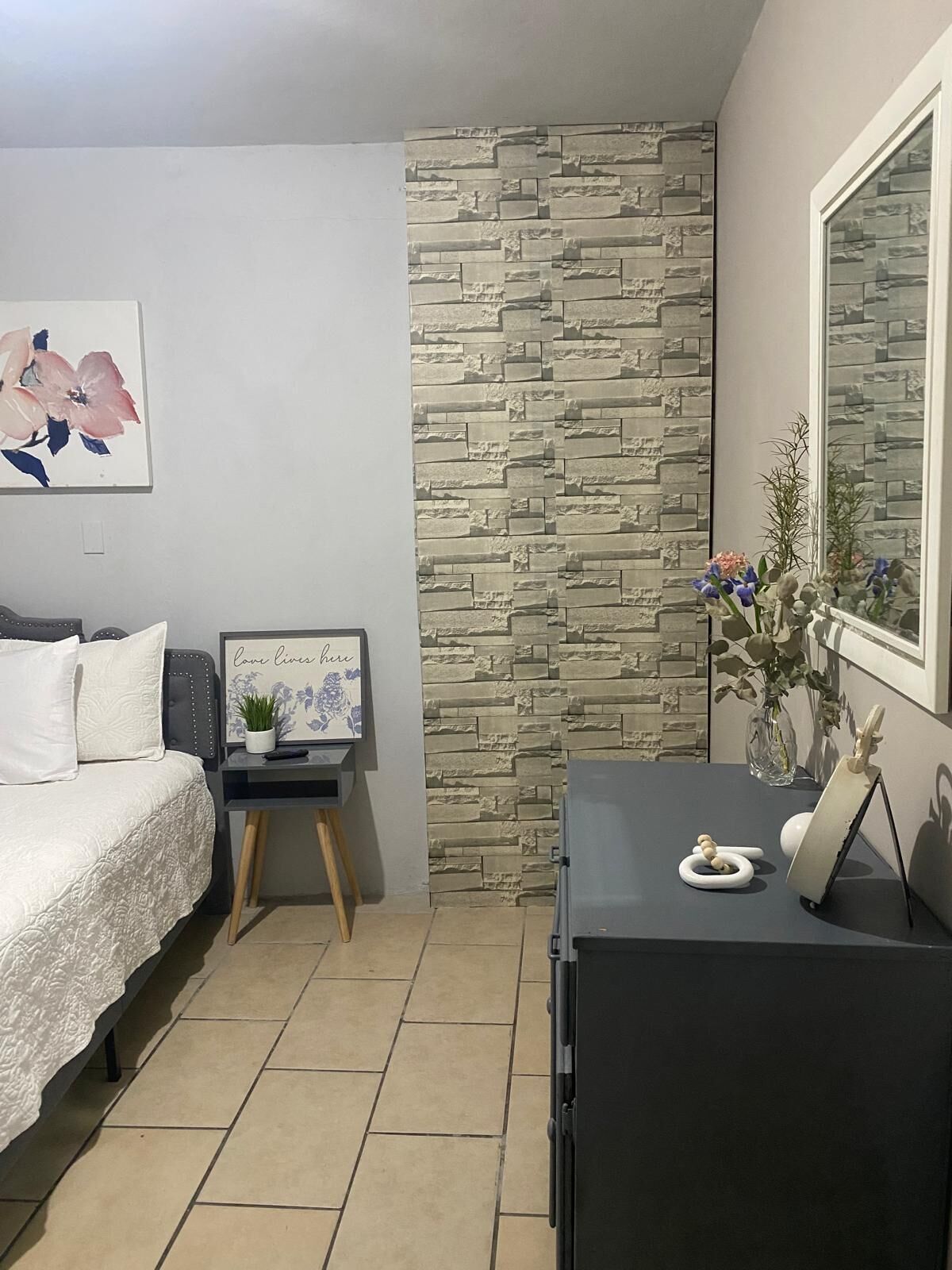 A bedroom with a bed, a small table with a plant, a mirror, and a wall with a stone-like texture.