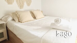 Un lit bien fait, avec du linge de lit blanc et des oreillers, une décoration murale en macramé et une serviette enroulée posée sur le lit.
