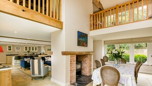 A spacious dining area with a brick fireplace, a high ceiling with exposed beams, and a view of the garden through large windows. - Stunning 4-Bedroom Barn Conversion (Sulgrave)