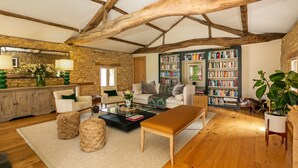 A spacious living room with exposed wooden beams, a fireplace, a large bookshelf, and a cozy seating area with a coffee table and cushions. - Stunning 4-Bedroom Barn Conversion (Sulgrave)