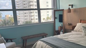 Une chambre à coucher comprenant un lit, une tête de lit en bois, une table de chevet, un banc, un bureau et une grande fenêtre donnant sur des immeubles et des palmiers.