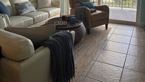 Un salon avec un canapé beige, une table basse et un fauteuil. On y trouve un ventilateur de plafond, une lampe, et une vue sur un balcon avec des immeubles en arrière-plan.