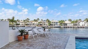 Un espace aménagé au bord de la piscine, avec des chaises longues et des plantes en pot, donnant sur une marina où sont amarrés des bateaux et où s’élèvent des palmiers.