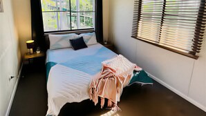 Un dormitorio con cama, ventilador de techo, ventana con persianas y mesita de noche con lámpara.