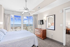 A hotel room with a bed, a dresser, a television, a ceiling fan, and a large window with a view of the city.