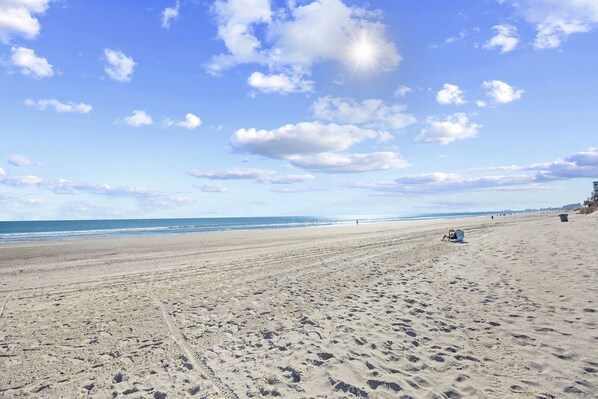 Una extensa playa arenosa con huellas, cielo despejado y edificios a lo lejos.
