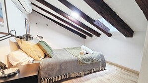 Ein gemütliches Schlafzimmer im Dachgeschoss mit schrägen Wänden, einem Bett mit gemustertem Überwurf, einem Schreibtisch mit Lampe und einer Wand-Klimaanlage.
