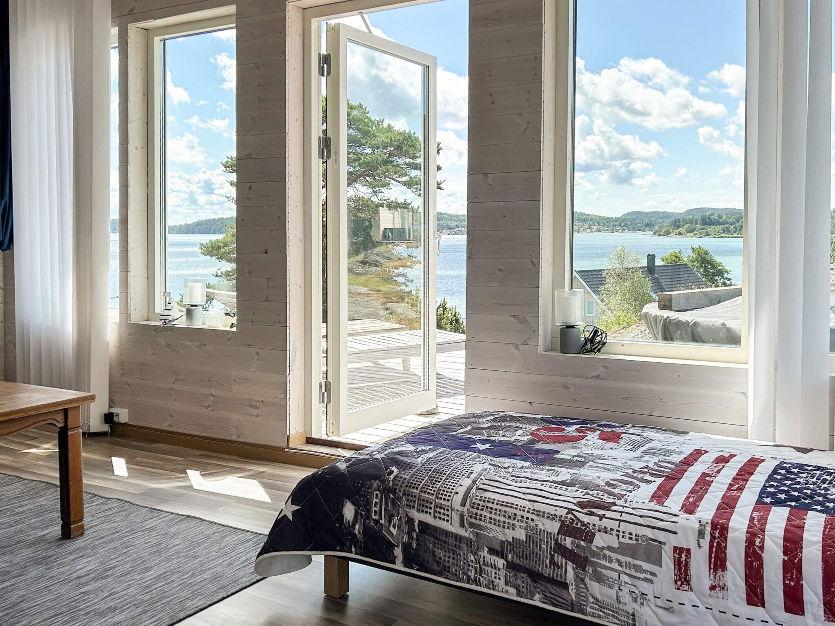 Une chambre avec une grande fenêtre, un lit recouvert d’une couvre-lit à motifs, et une vue sur un lac et des collines.