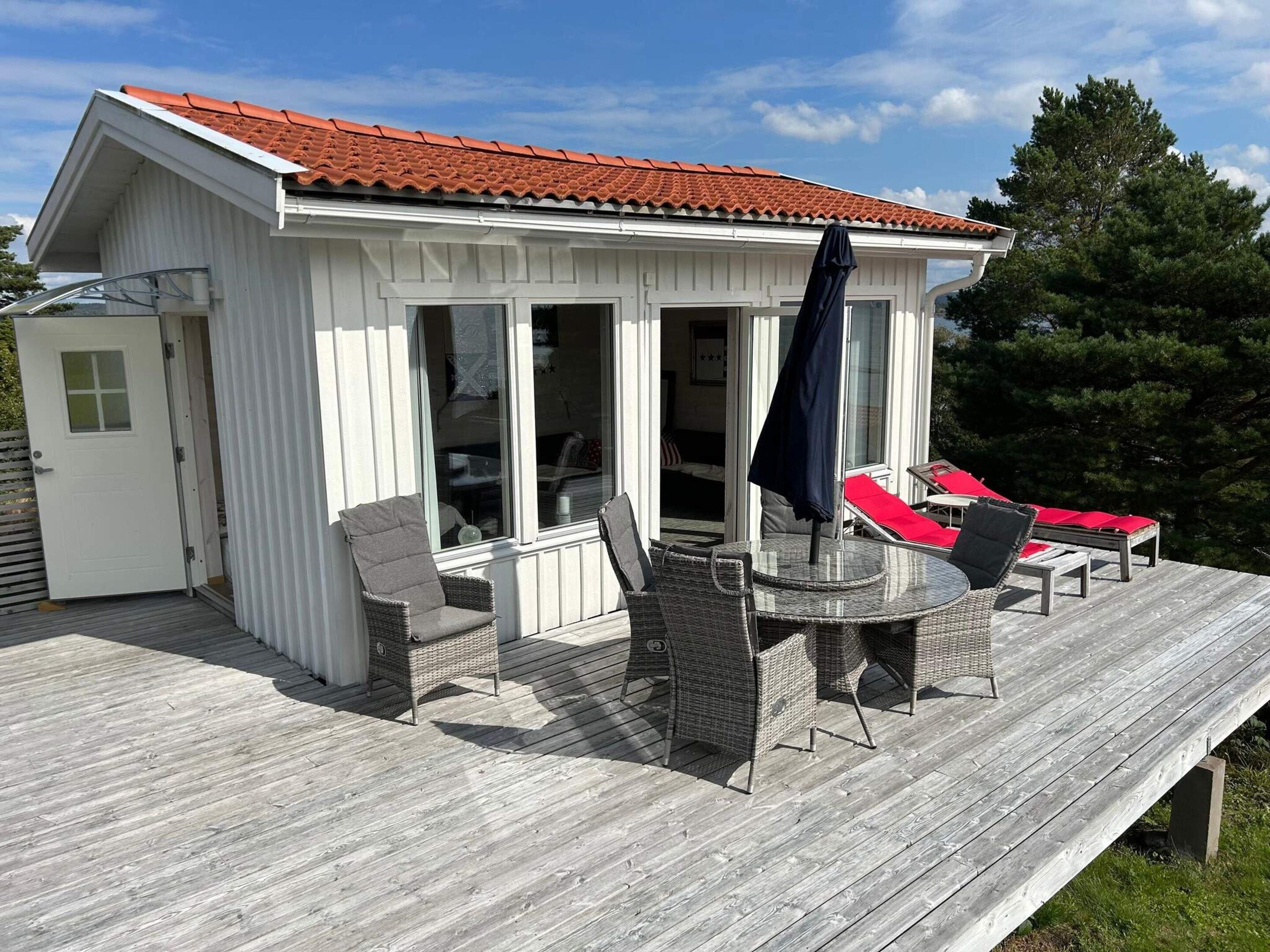 A covered outdoor seating area with wicker furniture and red cushions, a wooden deck, and a building with a red-tiled roof.