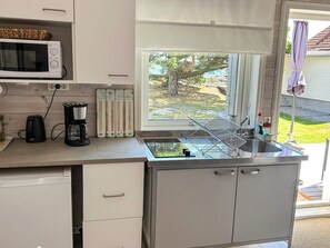 Eine kleine Küche mit einer Mikrowelle, einem Toaster und einer Kaffeemaschine auf der Arbeitsplatte, einem Fenster mit Blick auf Grünflächen und einem Spülbecken mit einem Geschirrgestell.