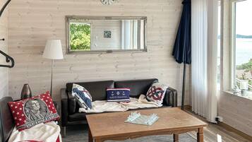 A room with a wooden ceiling, a sofa with patriotic pillows, a chessboard on a wooden table, and a view of the sea through a window.