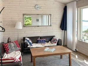 Ein Zimmer mit Holzboden, einer Couch mit patriotischen Kissen, einem Schachbrett auf einem Holztisch und Blick auf das Meer durch ein Fenster.