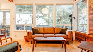 A cozy living room with a leather sofa, a coffee table, and a large window offering a view of trees. - Hot Tub + Forest Views | Mt Hood Getaway (Mount Hood Village)